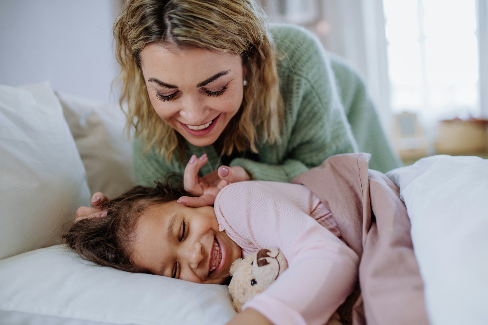 Woman smiling and cuddling a sleeping child on a bed with a teddy bear