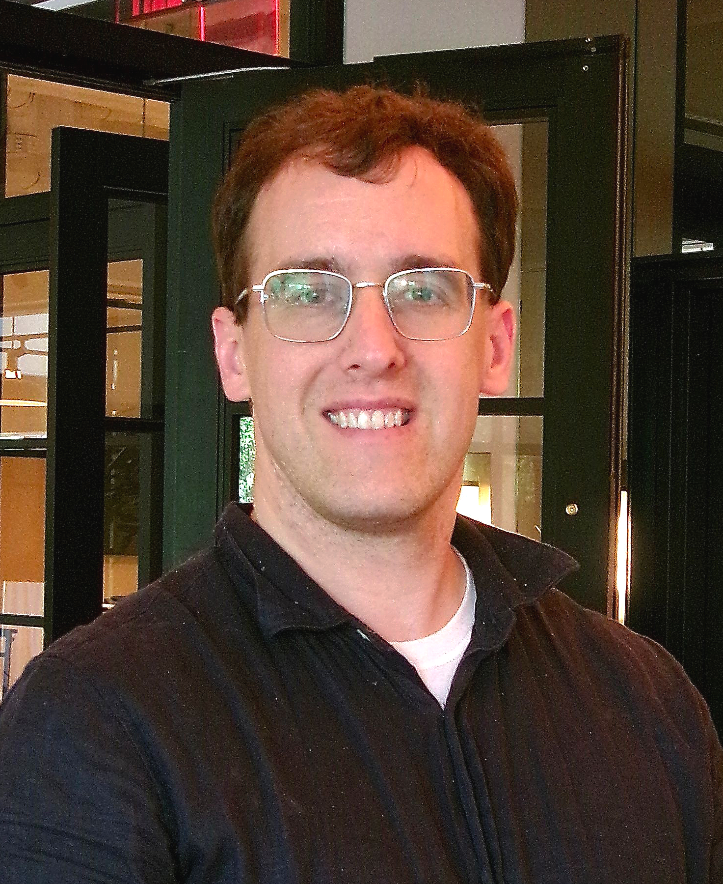 Dark haired man in black button up shirt and wire rimmed glasses smiles at camera.