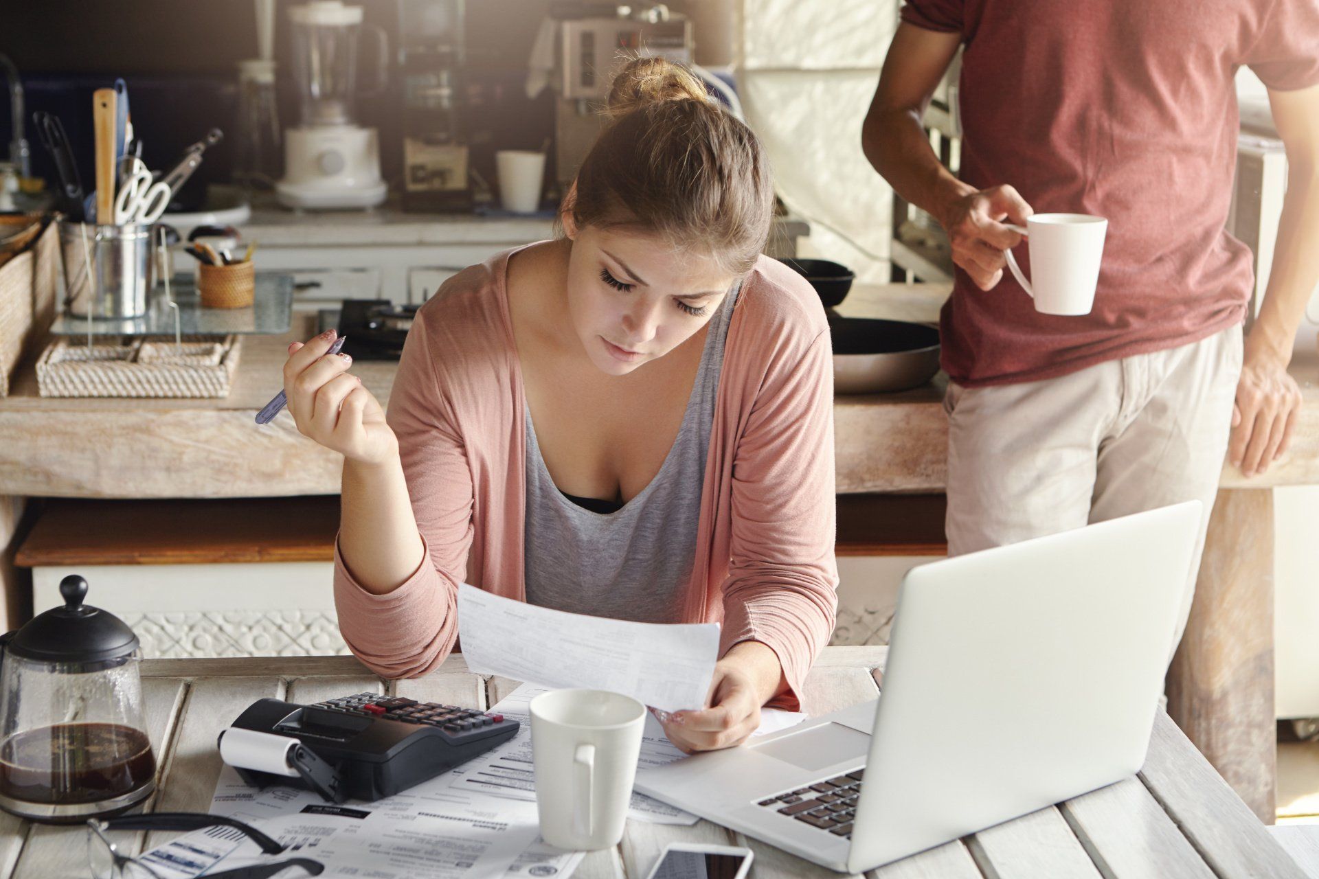 Woman reviews papers, calculating expenses at a table with laptop, man holding a mug in the background.