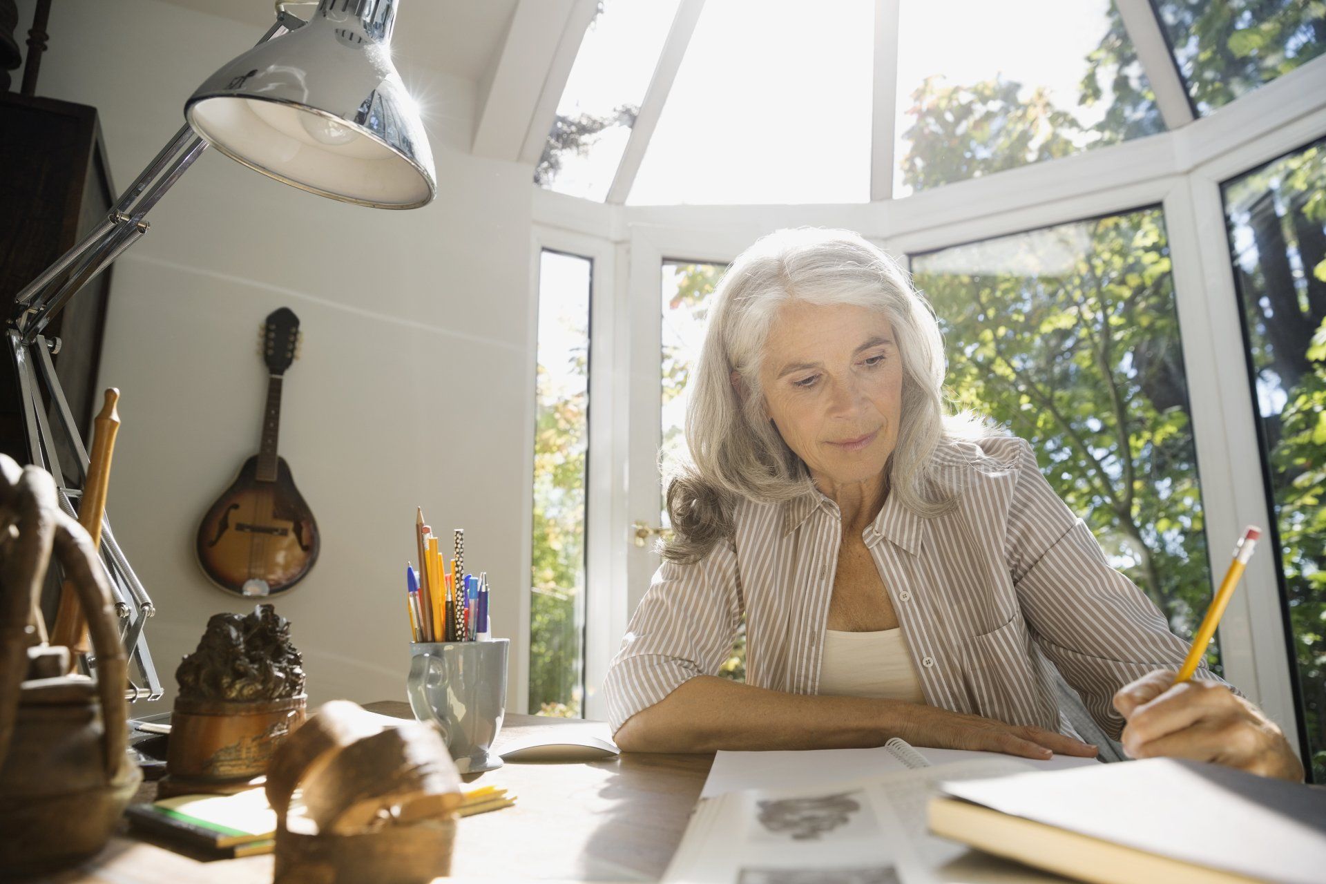 Woman with gray hair writing at a desk by a window. Sunlight streams in.