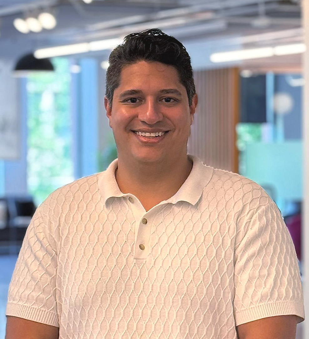 Dark haired man in white textured shirt smiles at camera in front of blurred office background.