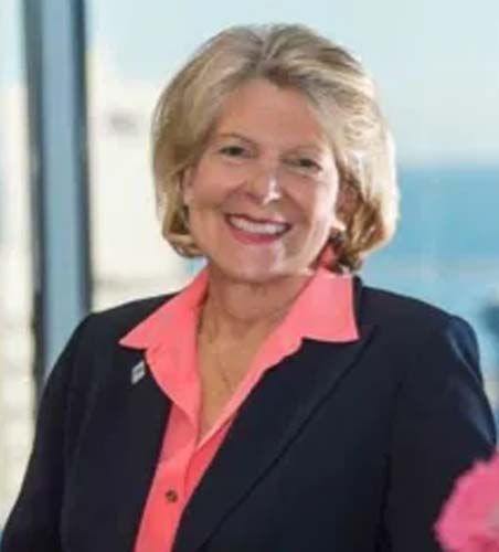 Woman in navy blazer and coral blouse smiles. Ocean view background.