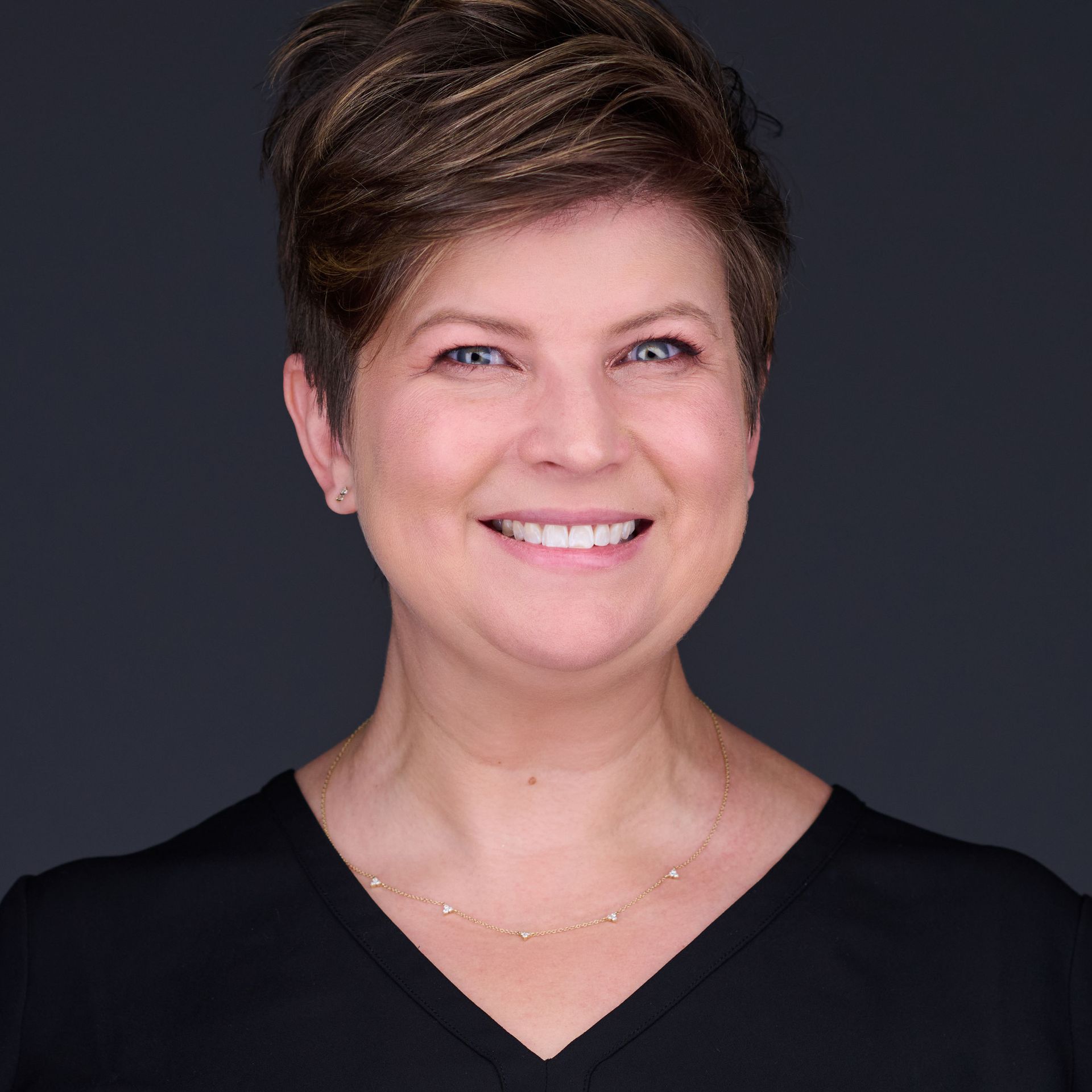 Smiling woman with short brown hair, wearing a black shirt and delicate gold necklace.
