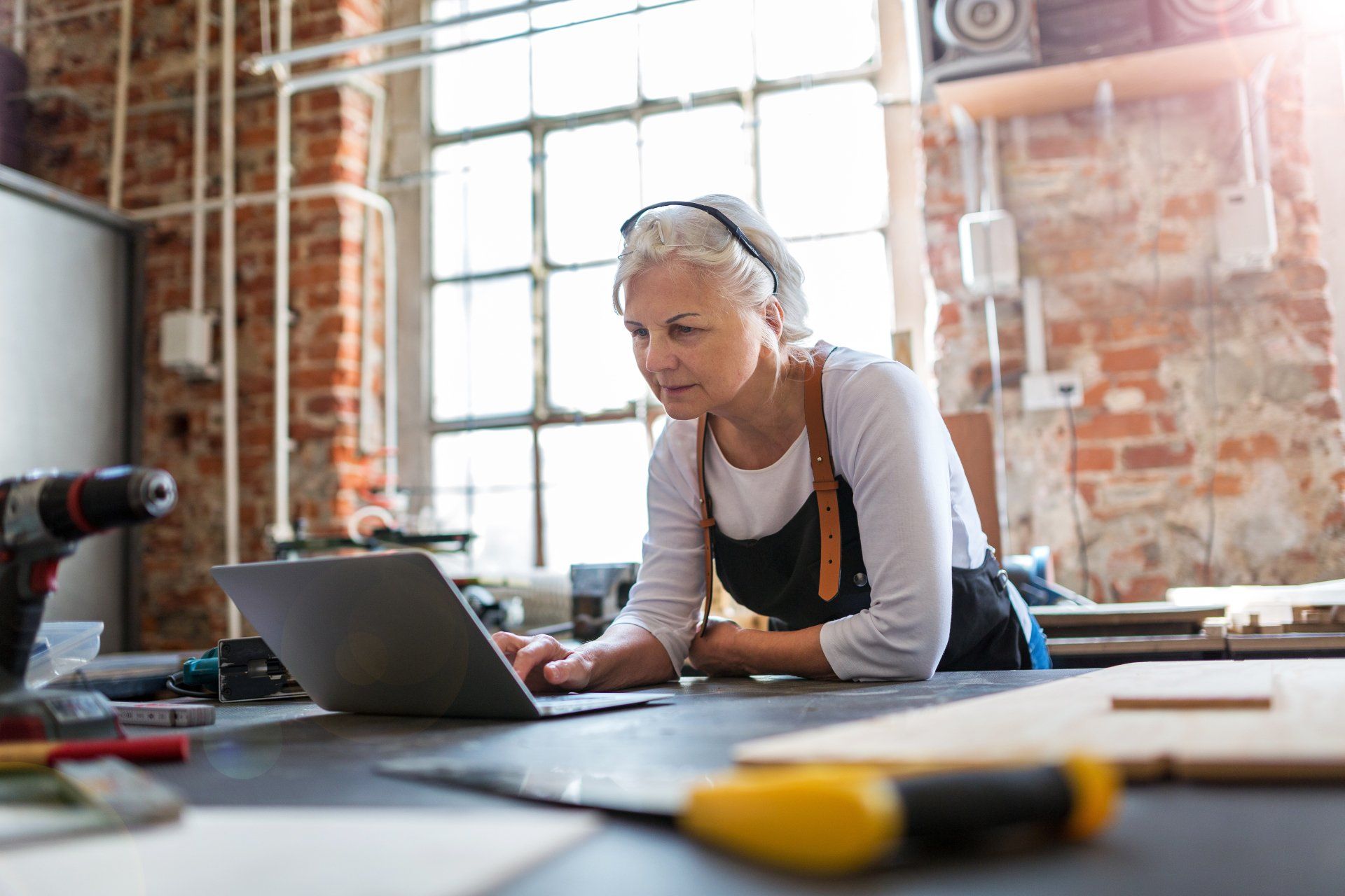 Woman in workshop using laptop at a workbench; tools and brick wall visible.