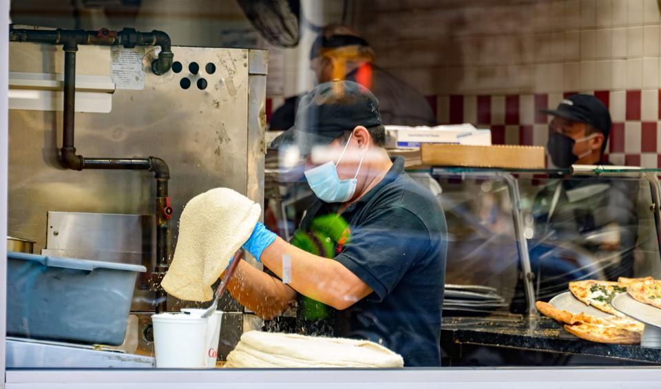 Chef in a kitchen wearing a mask, tossing pizza dough. Others in the background, pizza on display.