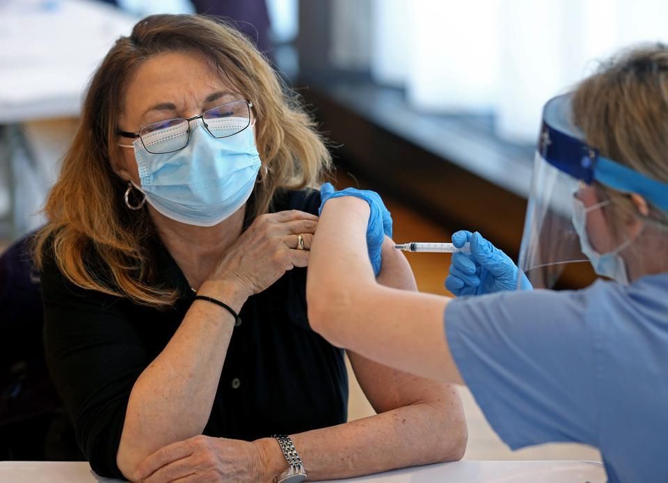 Woman wearing mask receiving a shot in her arm from a person wearing a face shield and gloves.