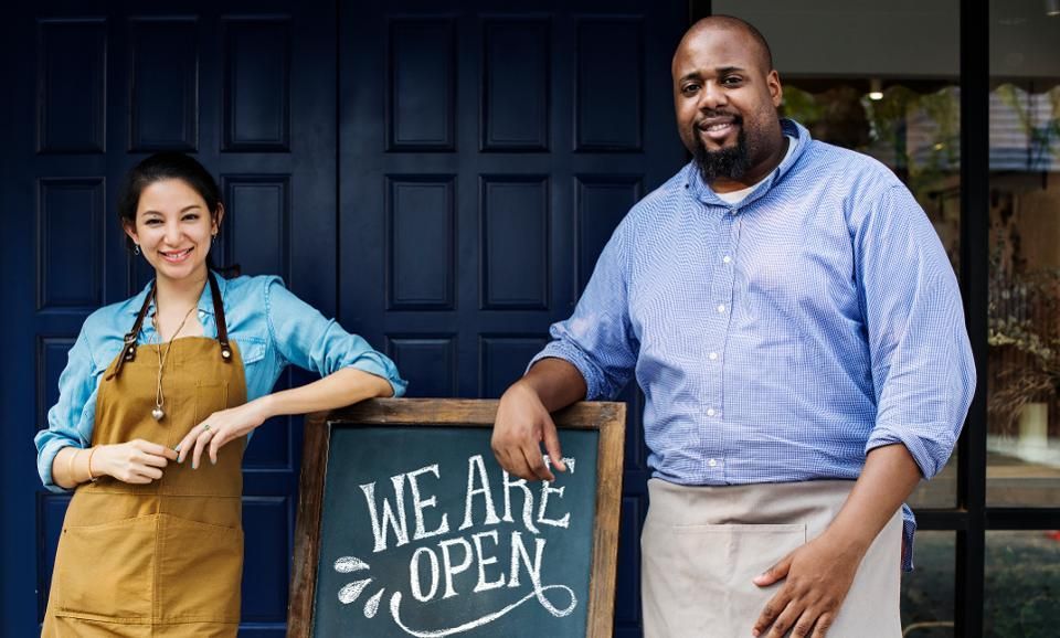 Two people wearing aprons stand by a chalkboard sign that says