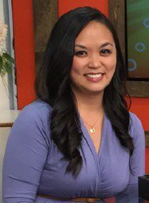 Woman with long, dark hair smiles. Wearing a lavender dress and gold necklace. Standing indoors with orange background.
