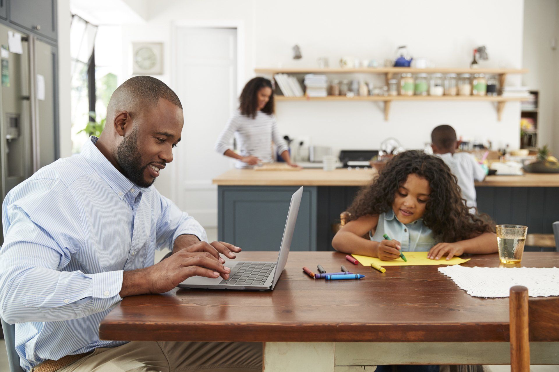 Man using laptop, girl drawing at a table in kitchen, woman cooking, and child in the background.
