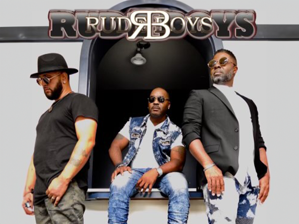 Three men are posing for a picture in front of a sign that says rude boys