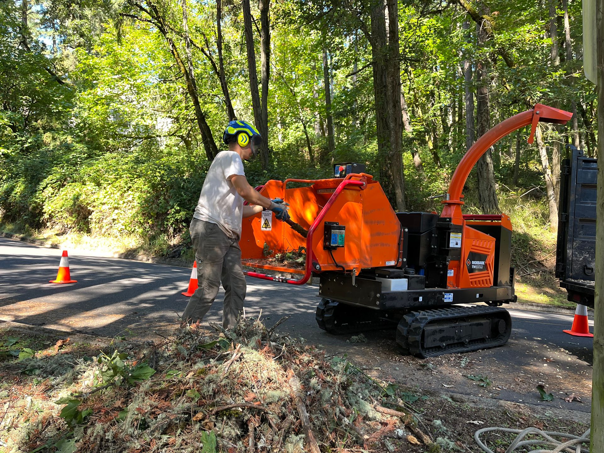 A person feeds tree branches into an orange wood chipper on a paved road.