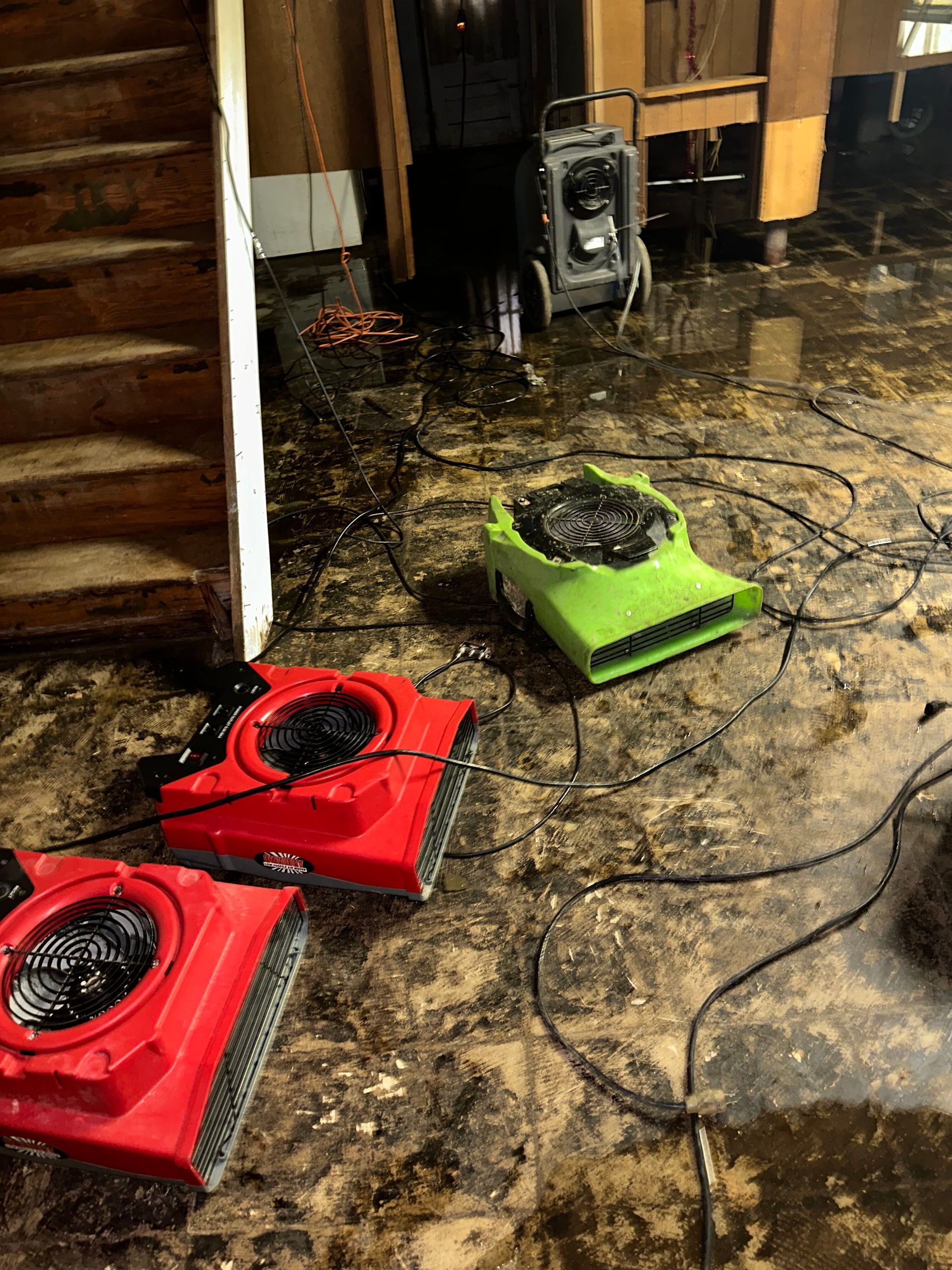 Red and green square floor fans on a wet, muddy floor near stairs and stacked speakers