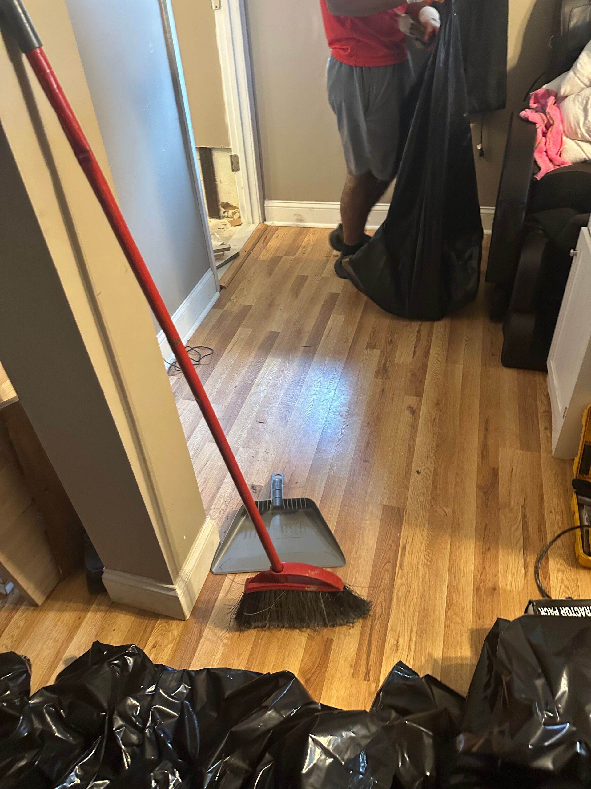 Person sweeping a hardwood hallway with a broom and dustpan beside black trash bags