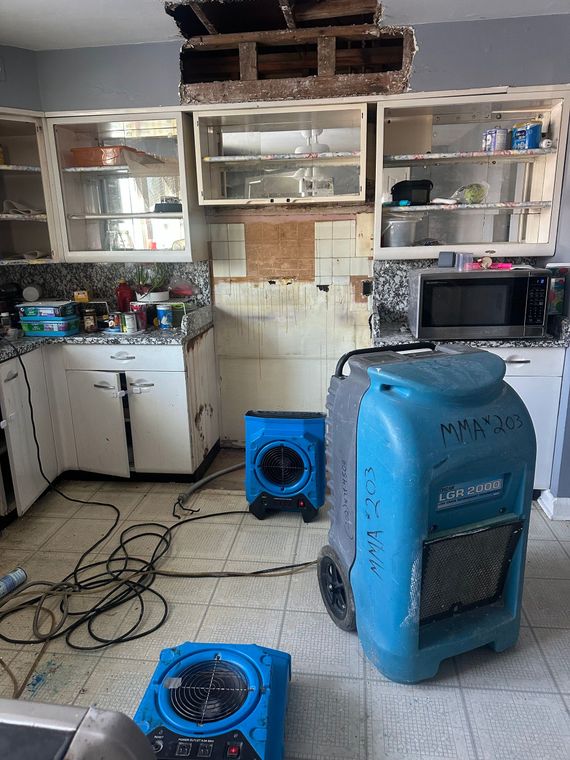 Kitchen with damaged ceiling and debris, two blue fans, and cords on the floor.