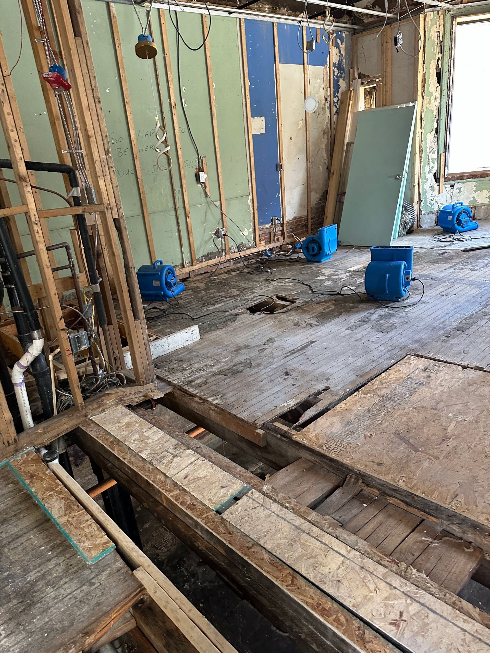 Construction site with exposed wooden floor joists, metal wall framing, and blue buckets on a concrete slab.