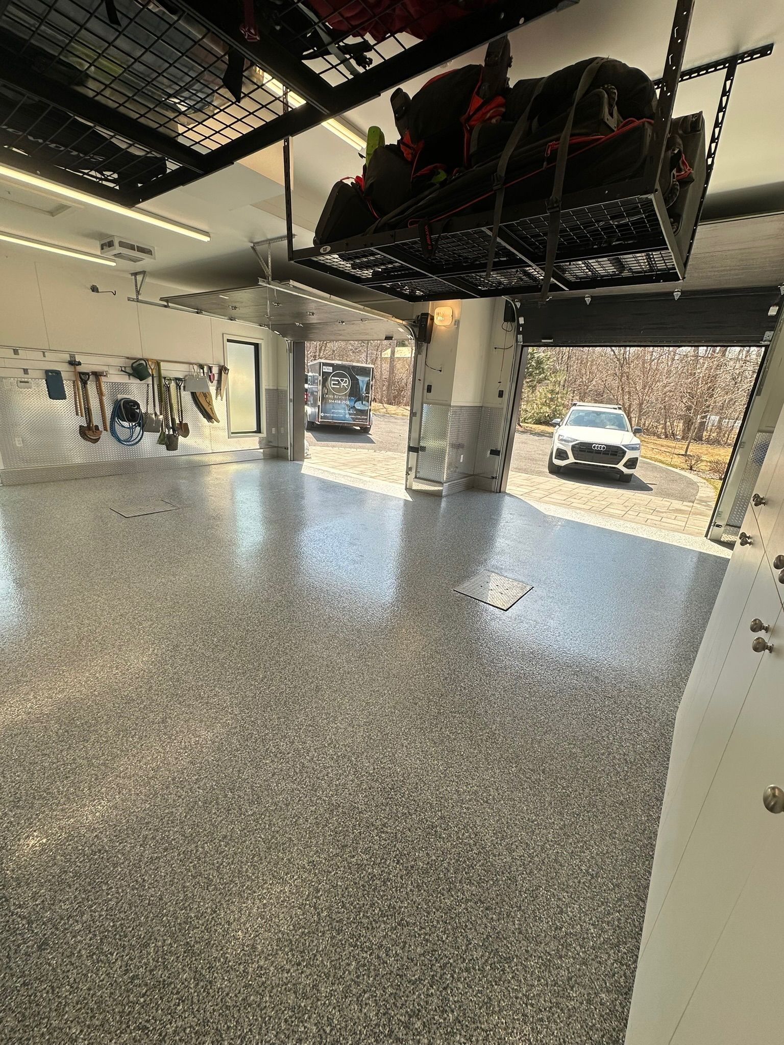 Garage spacieux avec sol en époxy moucheté, étagères de rangement suspendues et voitures garées devant la porte ouverte.