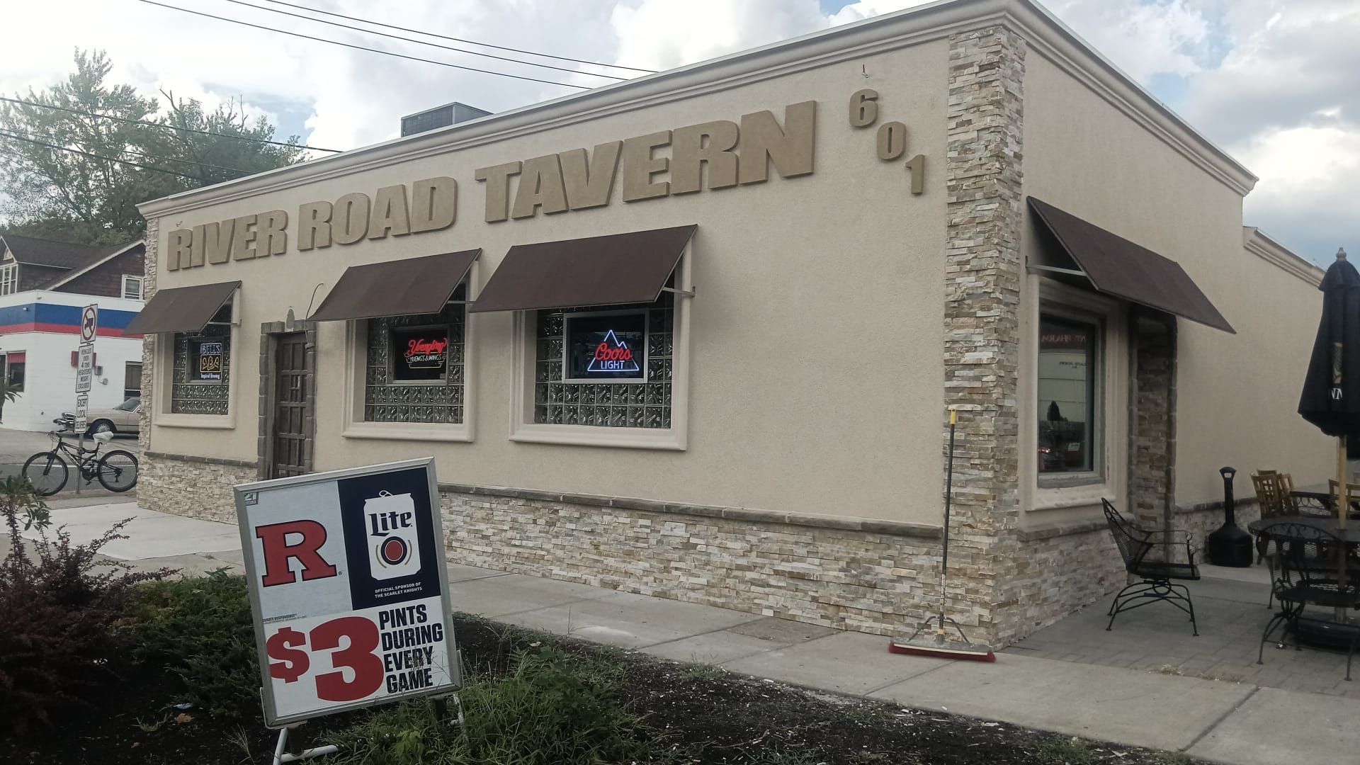 Exterior of a roadside tavern with a stone facade, neon window signs, and a $3 sign on the sidewalk