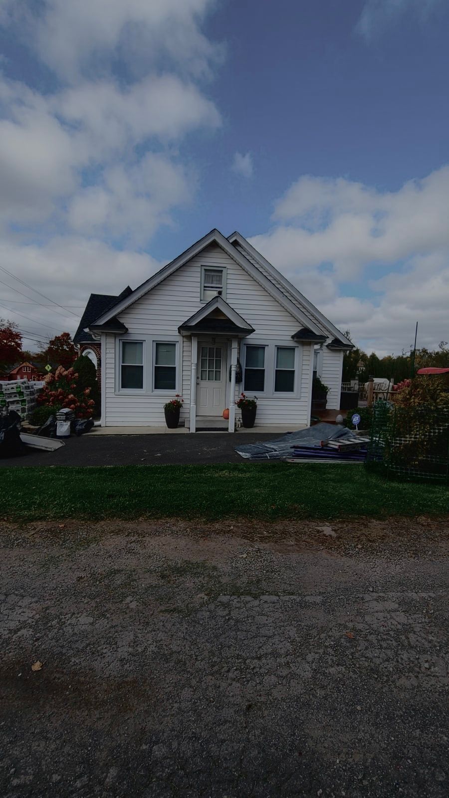 Small white house with porch and dark roof under a cloudy sky, seen from a gravel driveway