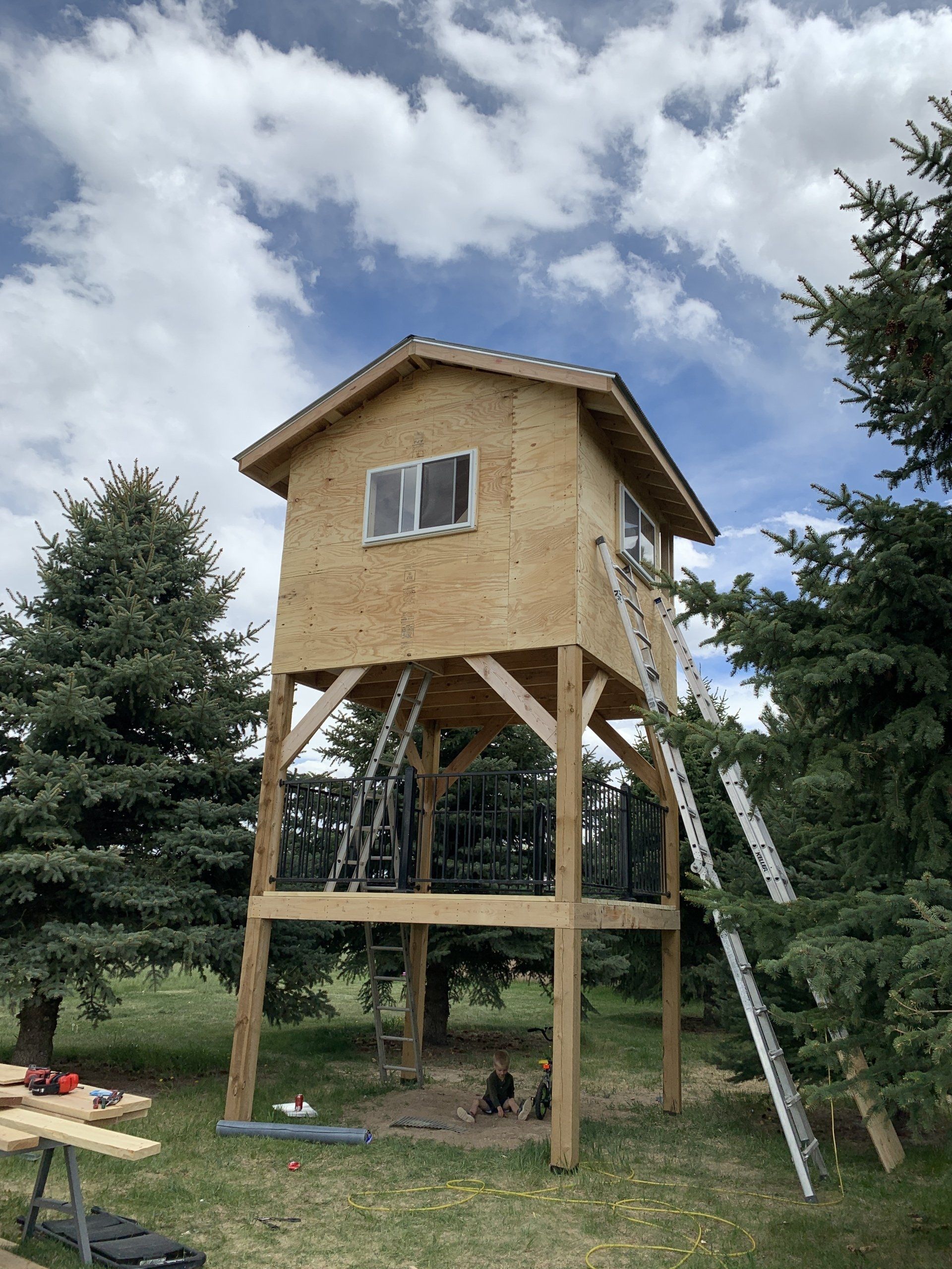 Wooden treehouse on stilts with ladder in a grassy yard under a cloudy sky