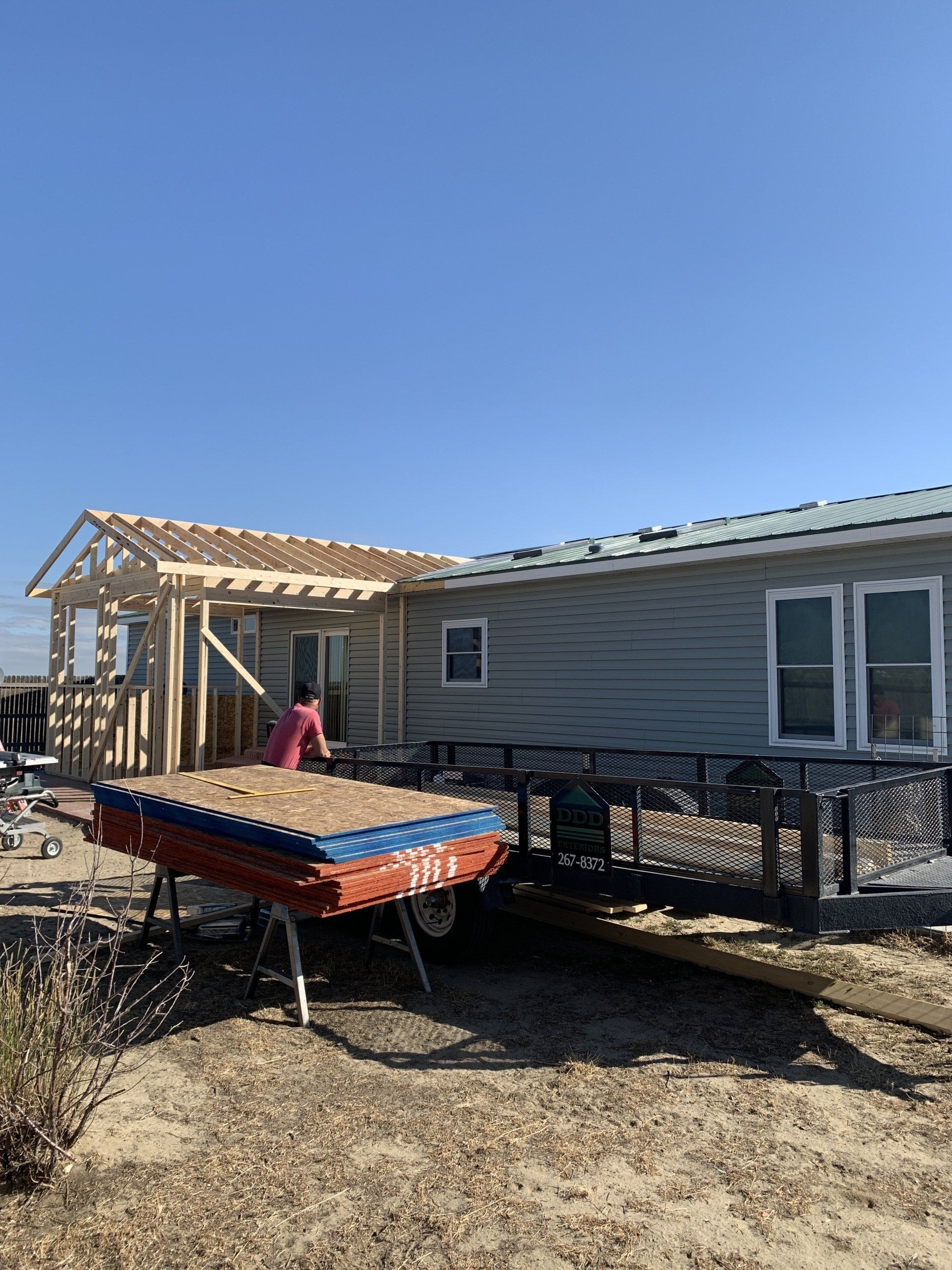 House under construction with a worker by a trailer in a dry yard, under a clear blue sky