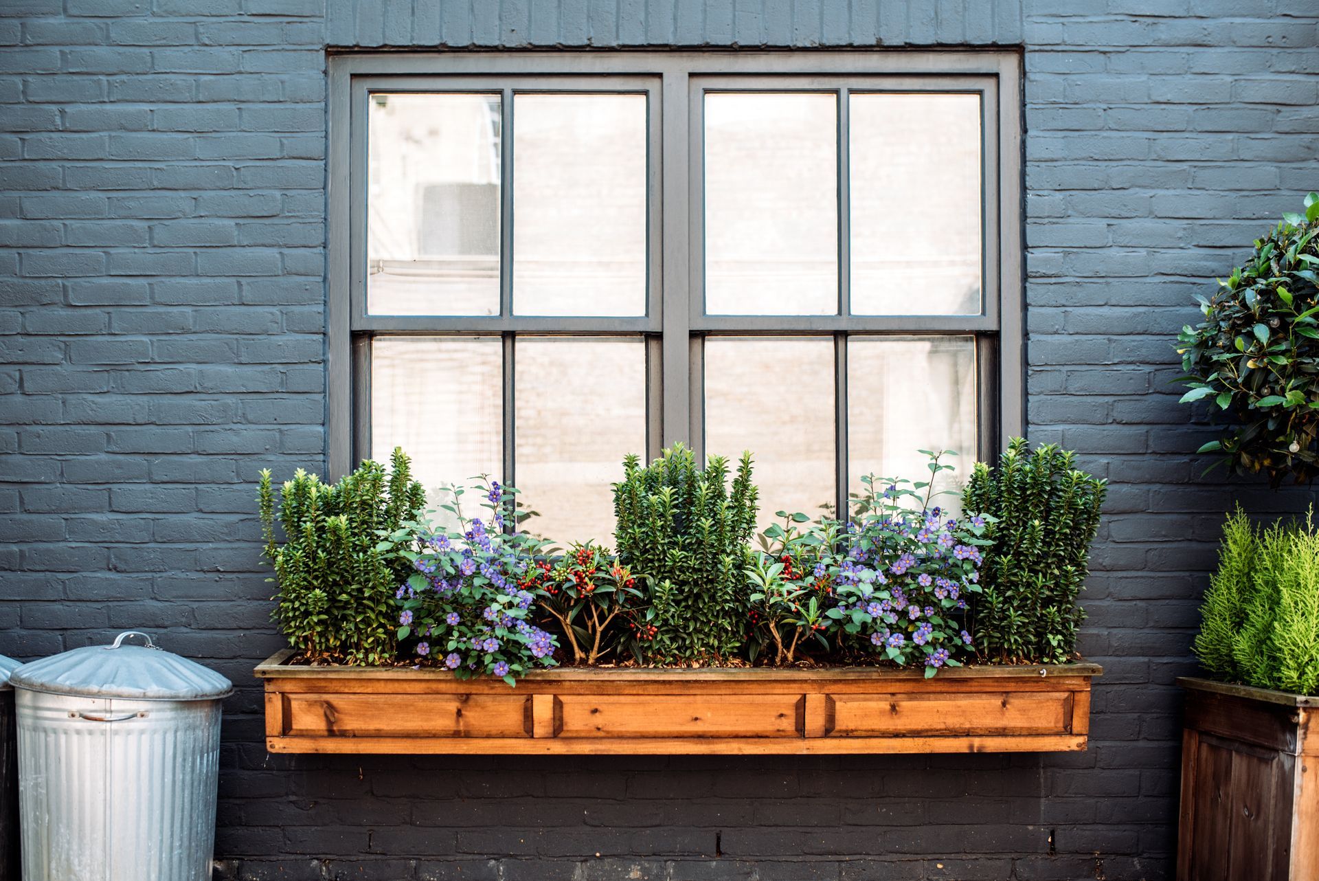 Blue brick wall with a window and a wooden planter box of flowers and greenery