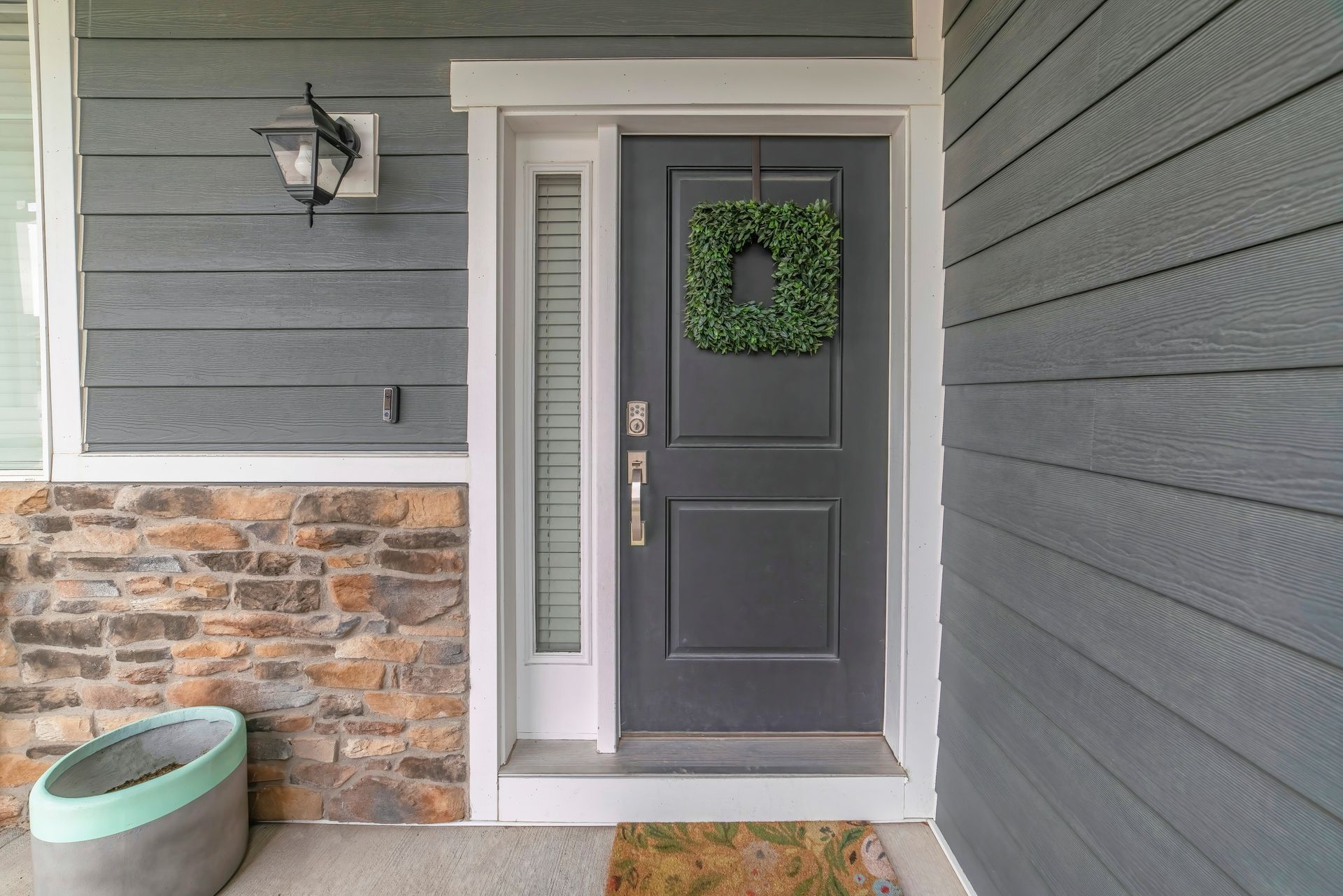 Front porch with gray door, green wreath, stone wall, and wall lantern beside a planter.