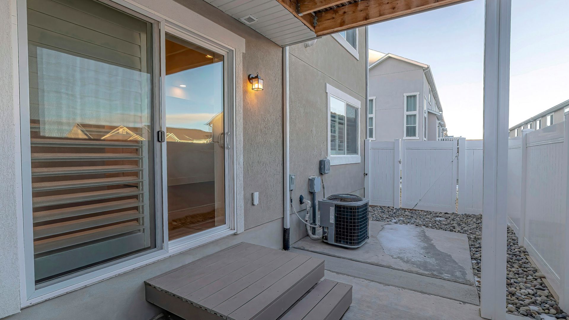 Patio with sliding glass door, bench, and HVAC units beside a beige townhouse at sunset