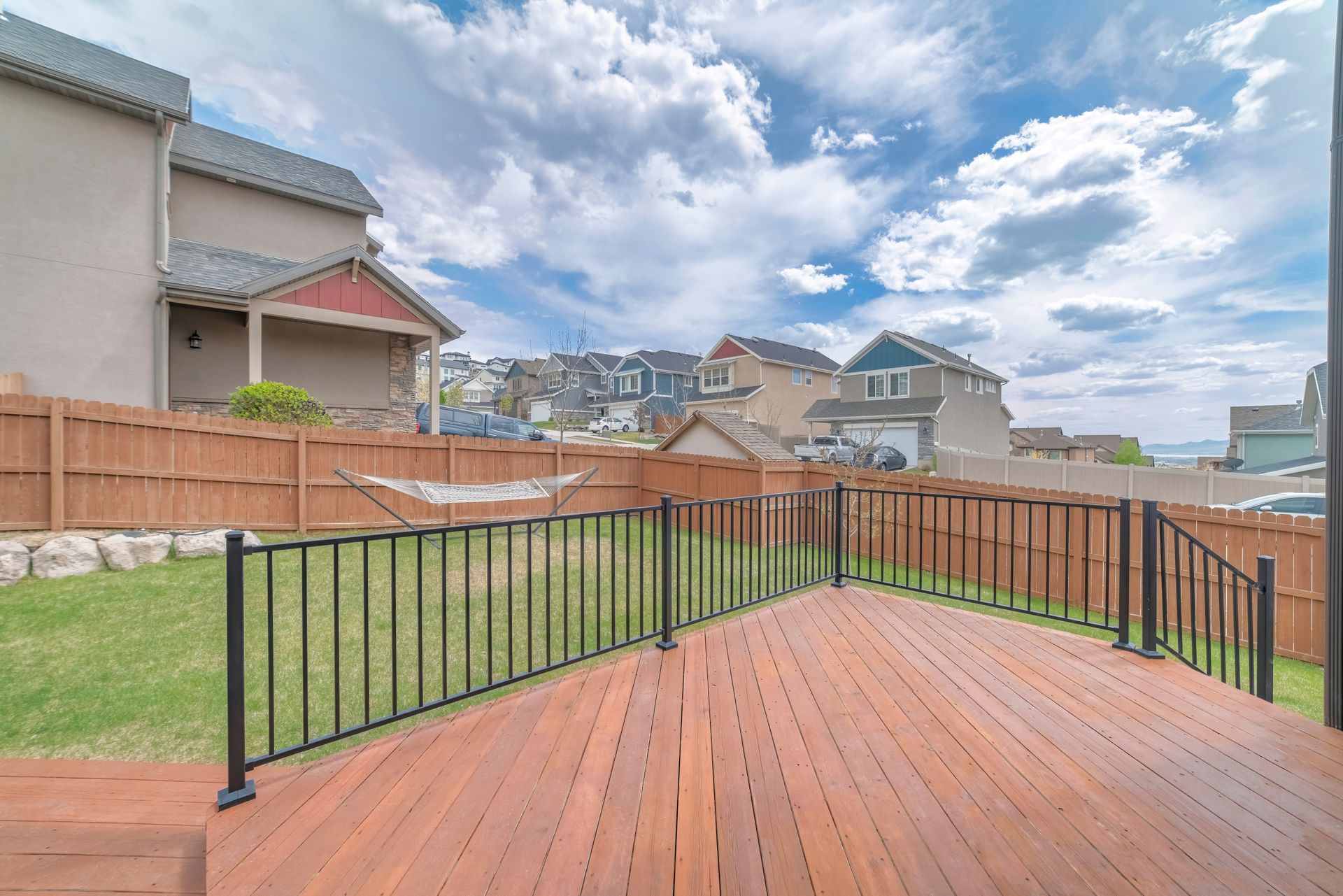 Wooden deck with black railing overlooking a grassy backyard and suburban houses under a cloudy sky