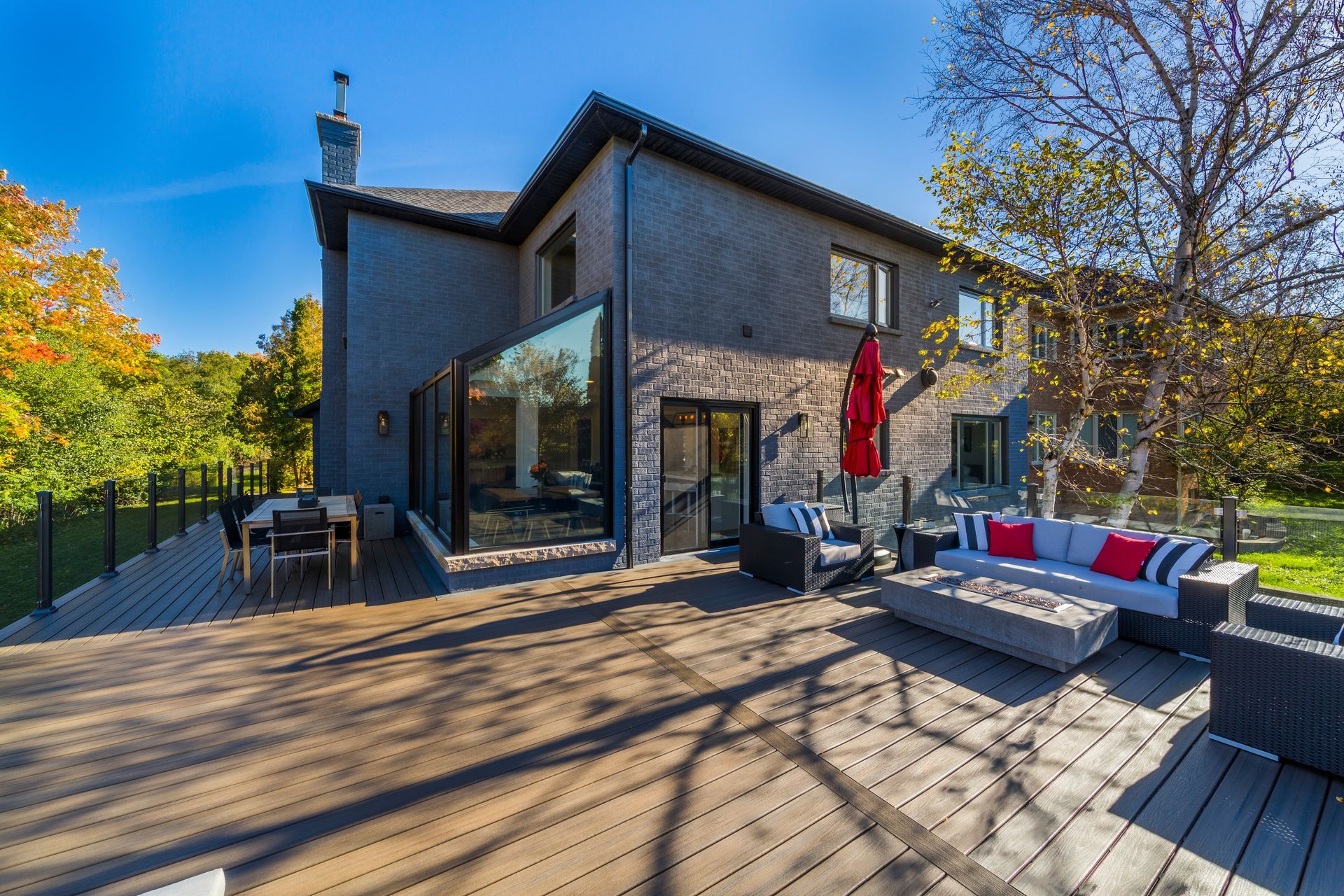 Modern house with a wooden deck, outdoor seating, red umbrella, and blue sky surrounded by trees Modern house with a wooden deck, outdoor seating, red umbrella, and blue sky surrounded by trees