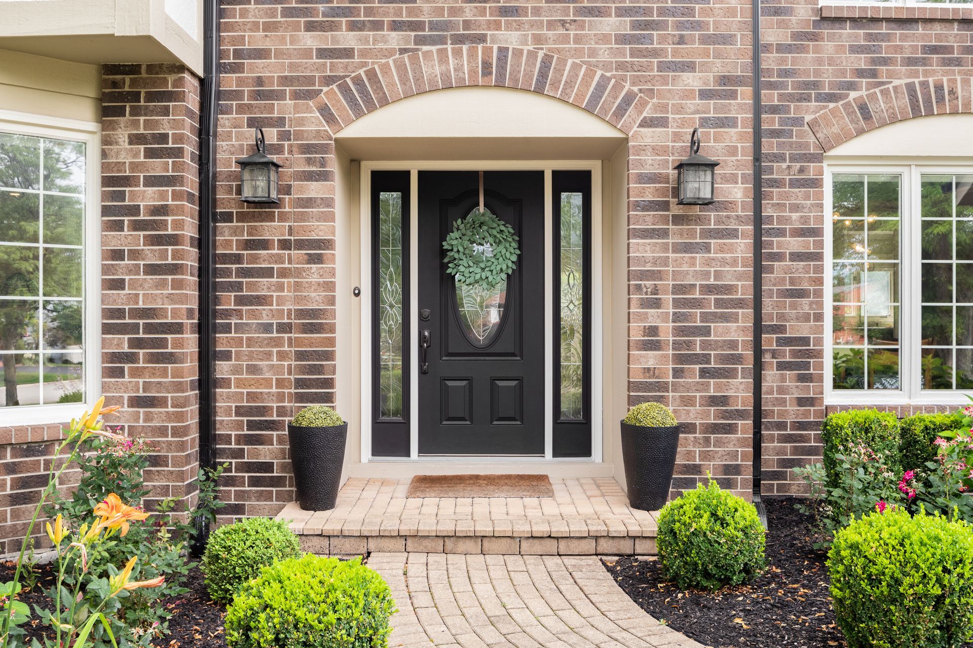 Brick house front entrance with black door, arched trim, potted plants, and landscaped walkway