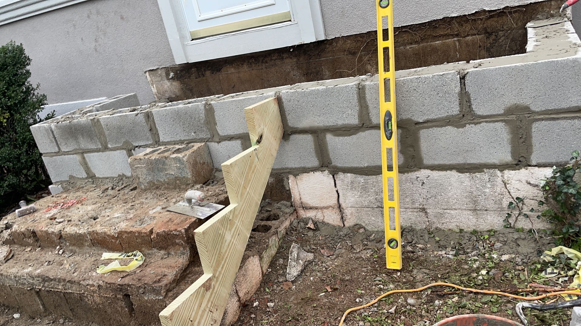 Partially built concrete block retaining wall beside house steps, with a yellow measuring tape in front