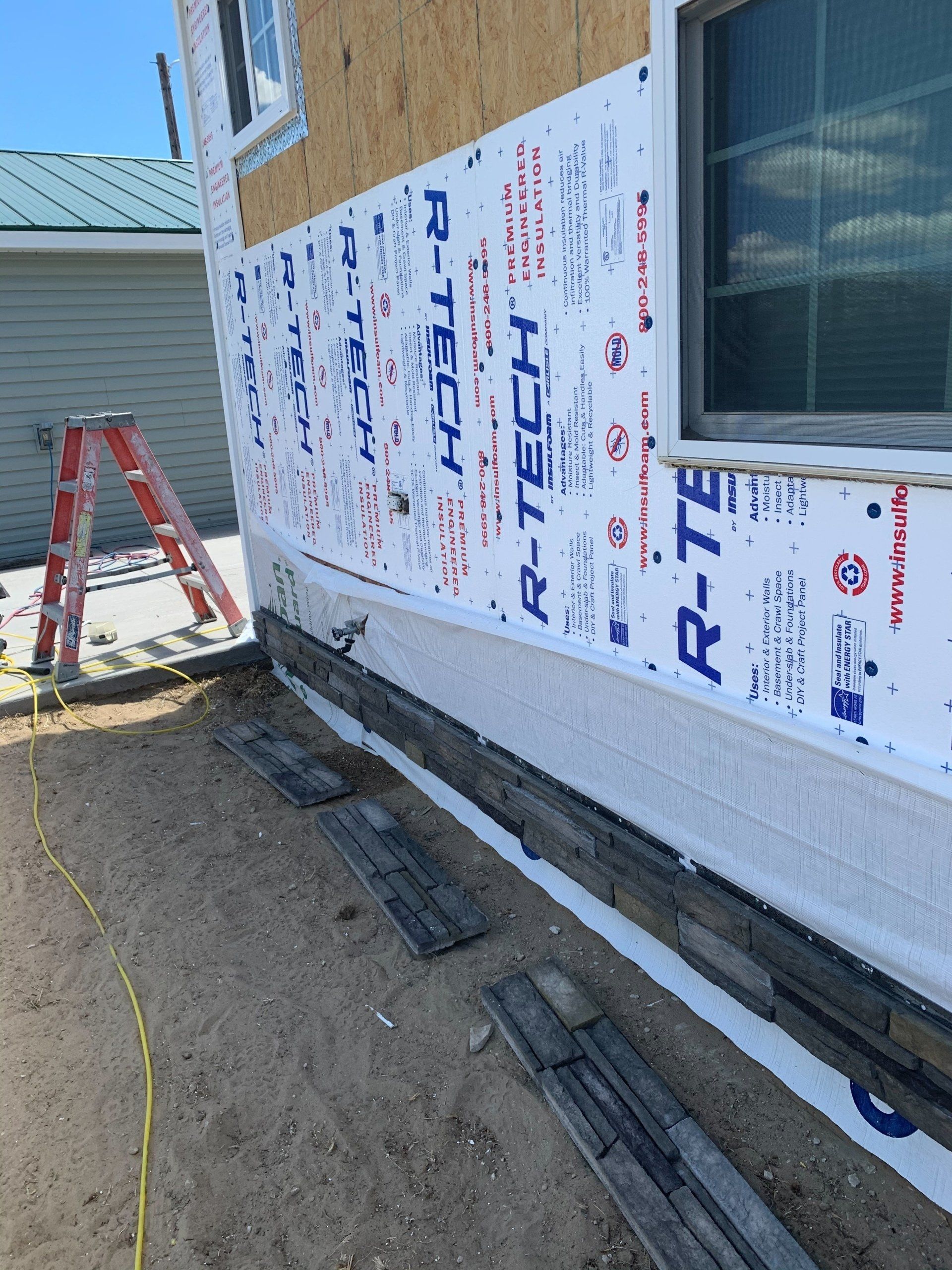 Partially finished house exterior with Tyvek wrap, window, ladder, and construction materials beside dirt ground