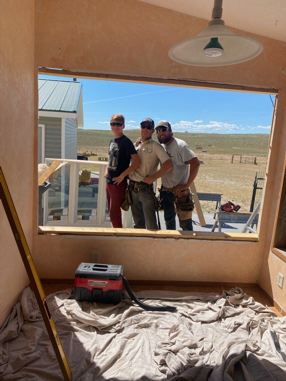 Three workers outside a window opening in a partially finished room with a tarp-covered floor.