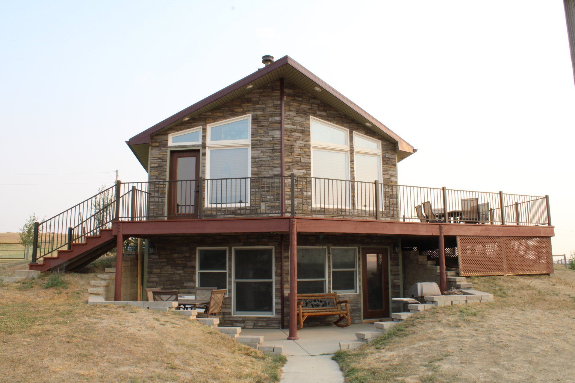 Two-story wooden house with a raised deck and stairs in a grassy, rural setting.