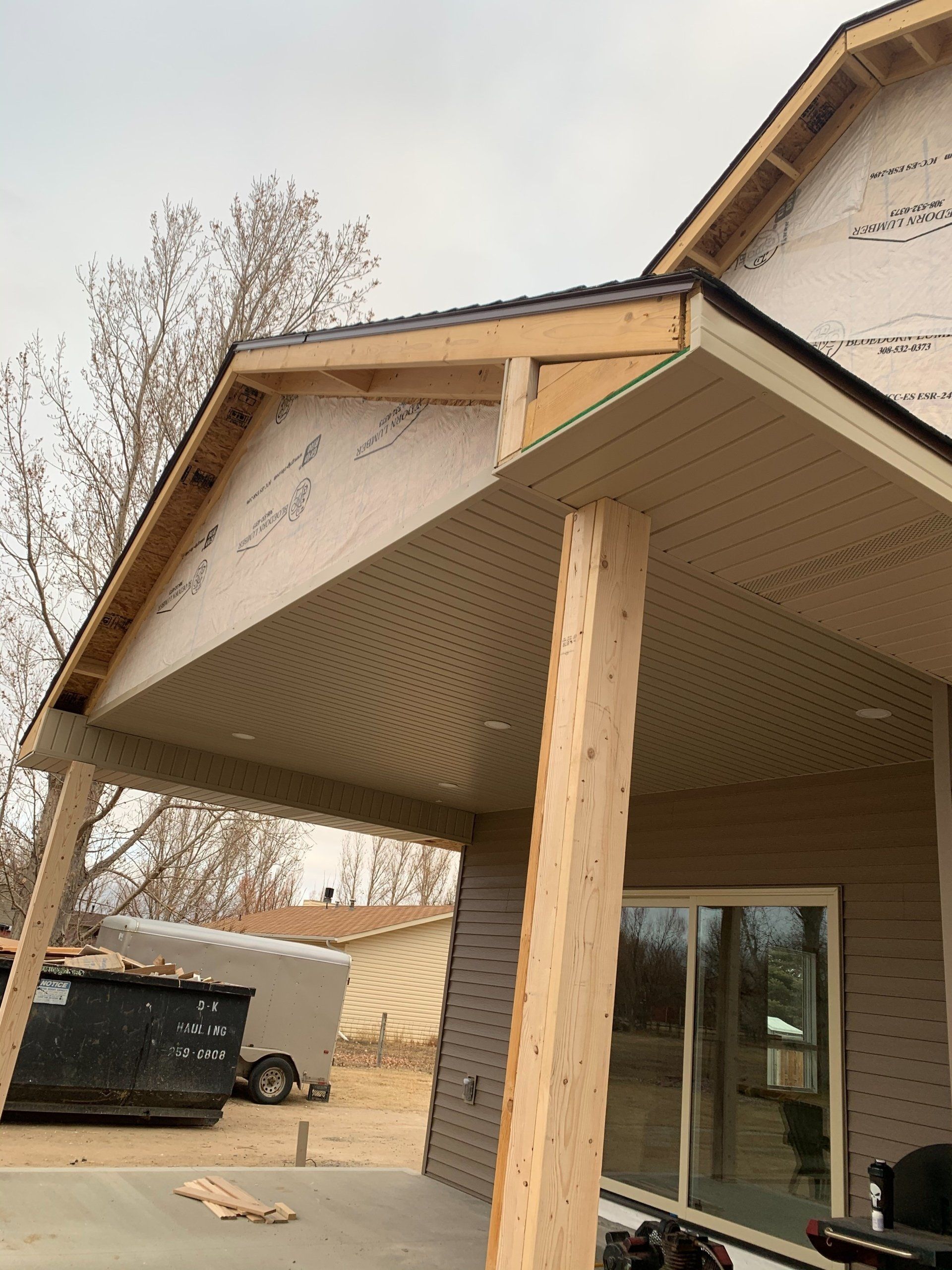Wooden porch roof under construction beside a beige house, with a support post and exposed plywood beams.