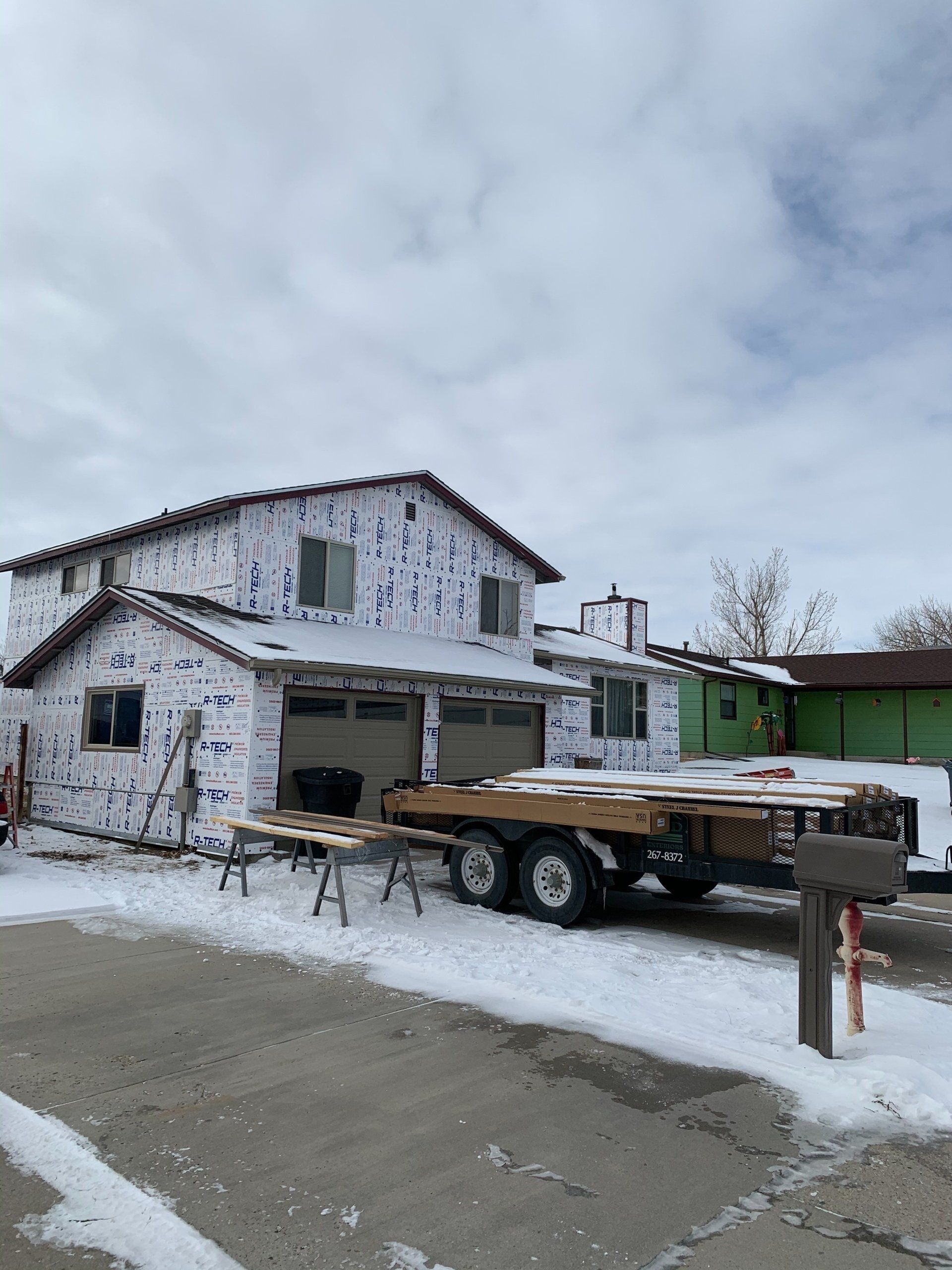 Snow-covered white building with green shed and a flatbed trailer parked in front under a cloudy sky