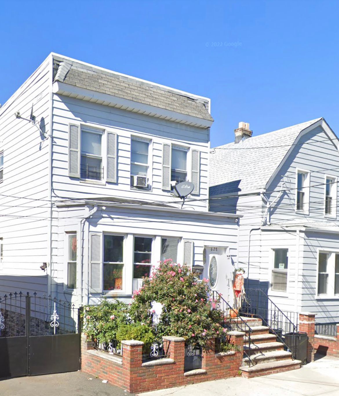 White two-story house with front steps, small garden, and a neighboring home under a bright sky