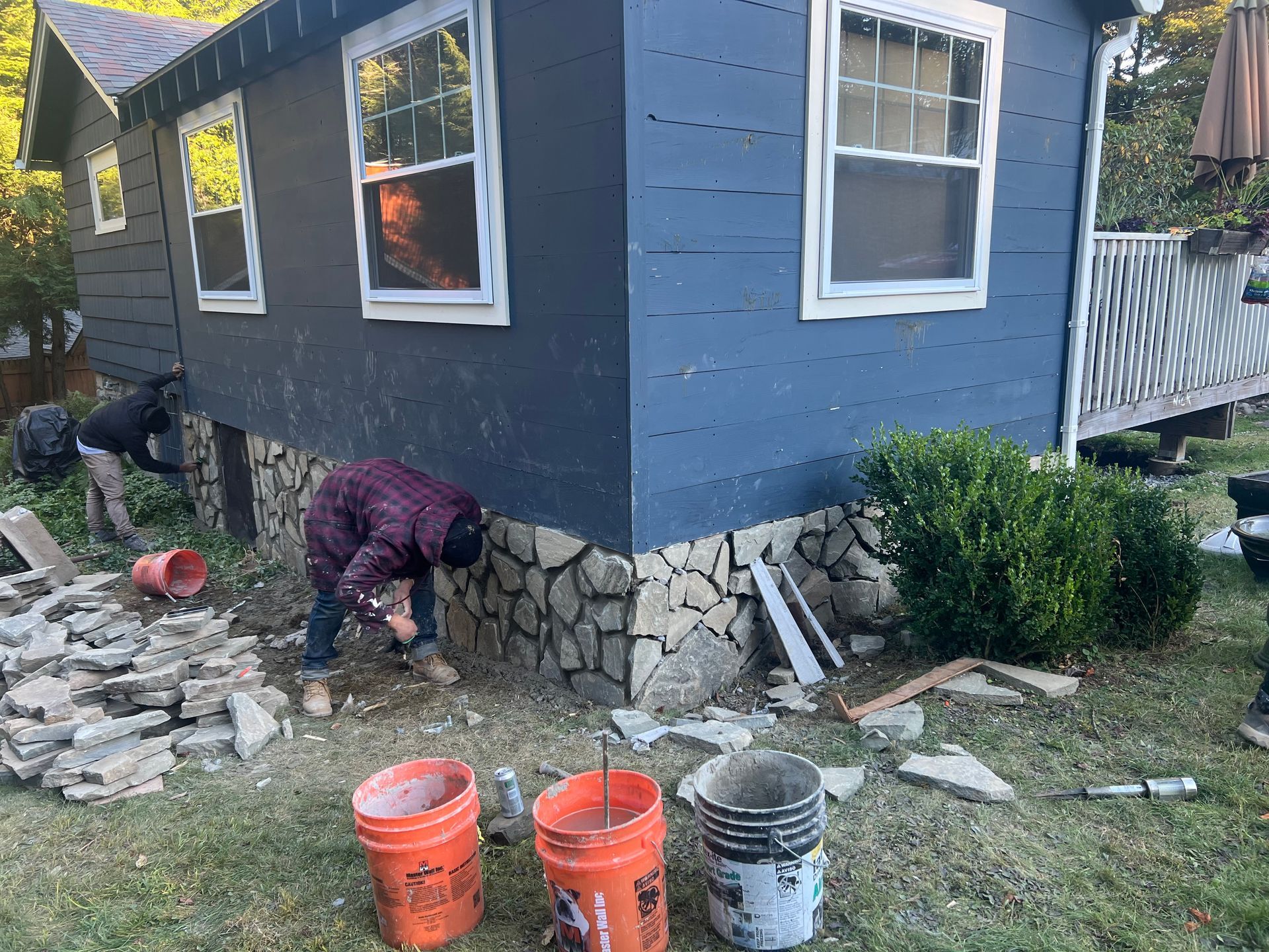 Person painting a blue house exterior with stone foundation and buckets on the ground