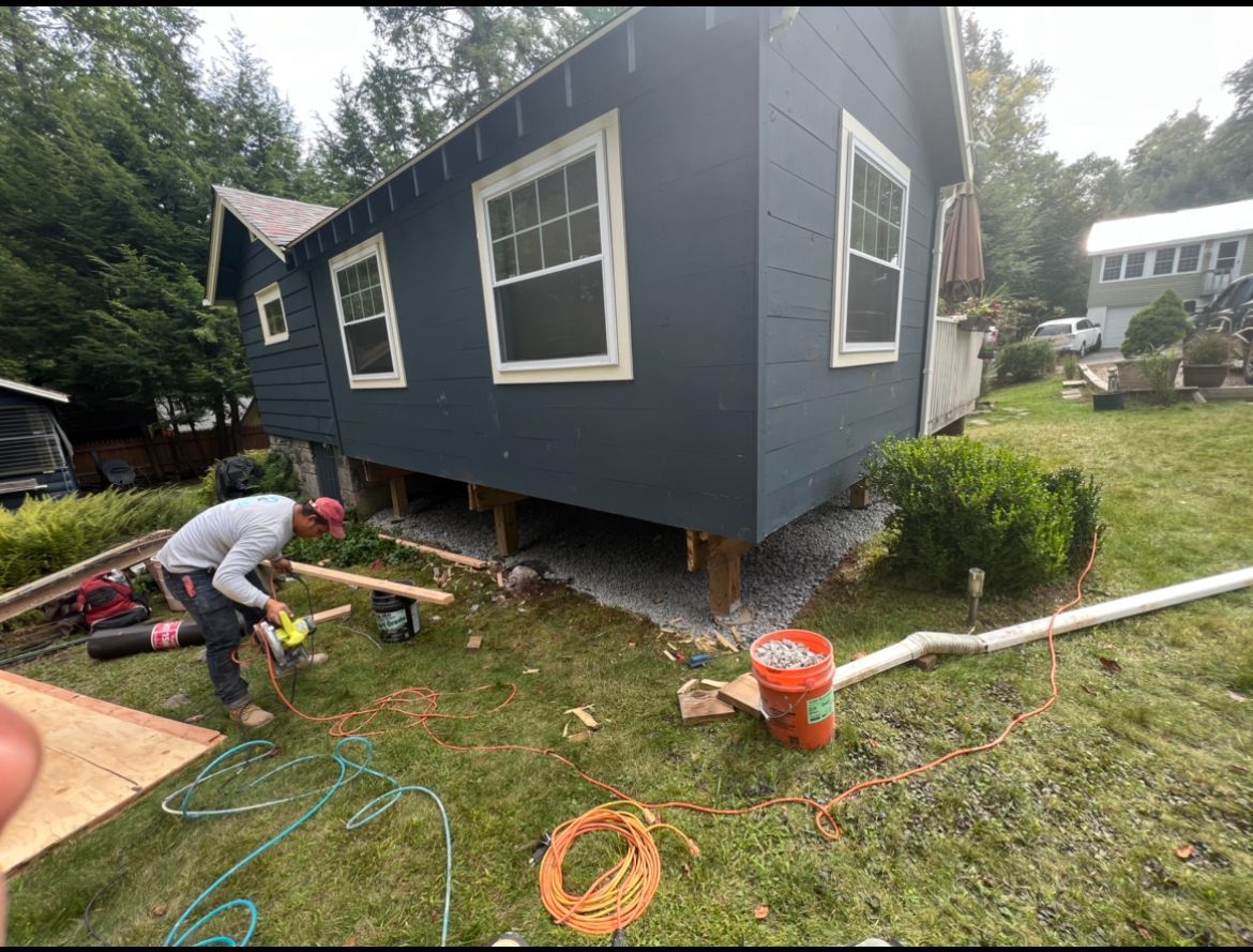 Worker repairing pipes under a raised gray house in a grassy yard, with orange hose and tools nearby