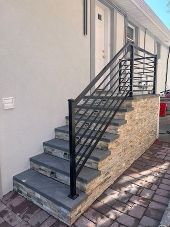 Exterior staircase with black metal railing beside a light-colored building and cobblestone walkway