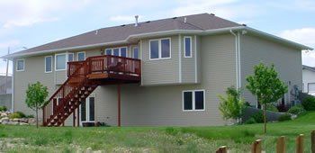 Suburban two-story beige house with a red wooden deck and stairs, surrounded by grass and trees