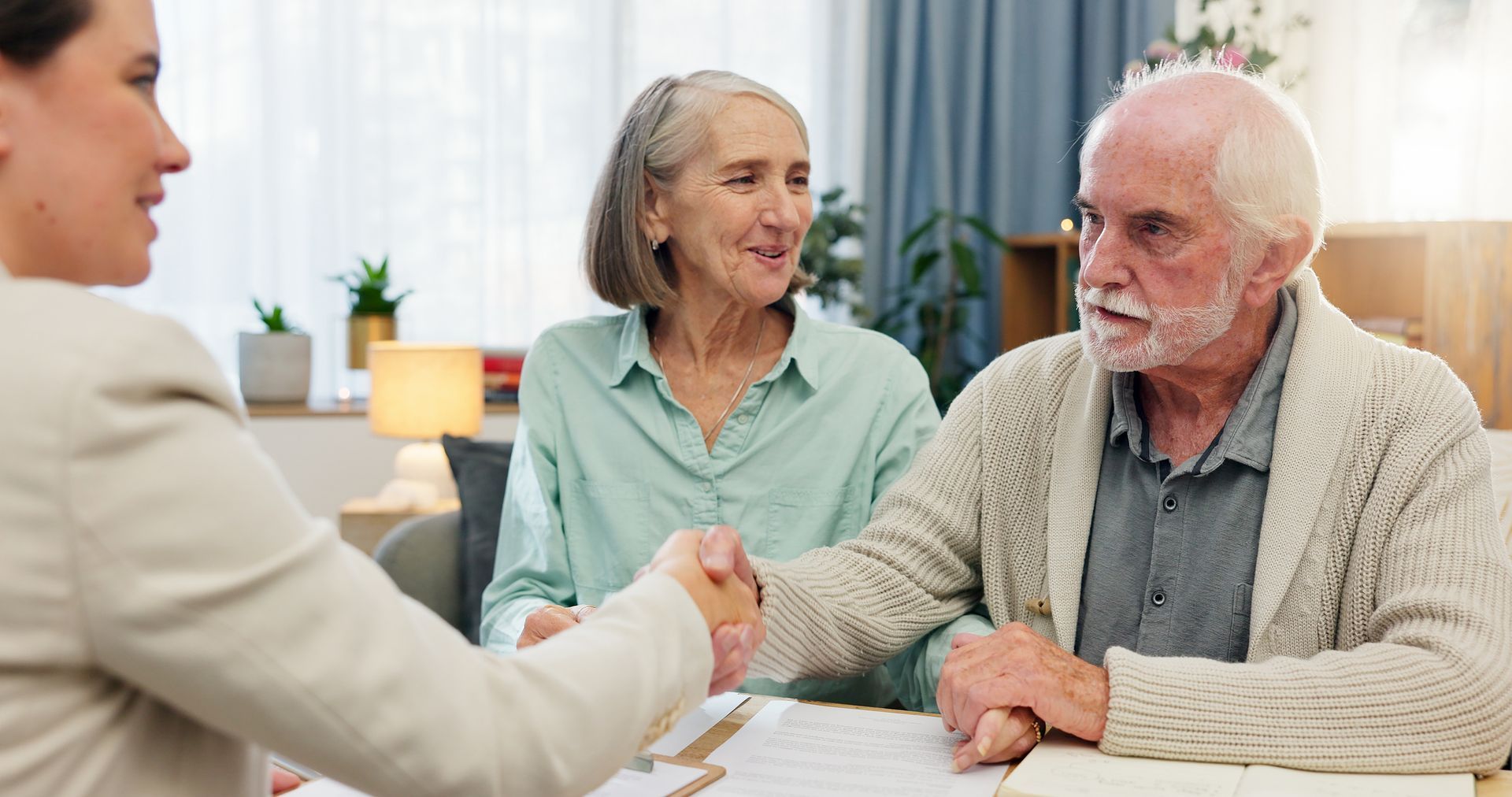 A young woman shakes hands with an elderly man seated with his wife at a table; sunlight streams in.