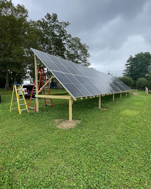 Solar panel array on metal frames in a grassy park under cloudy skies, with a ladder nearby.