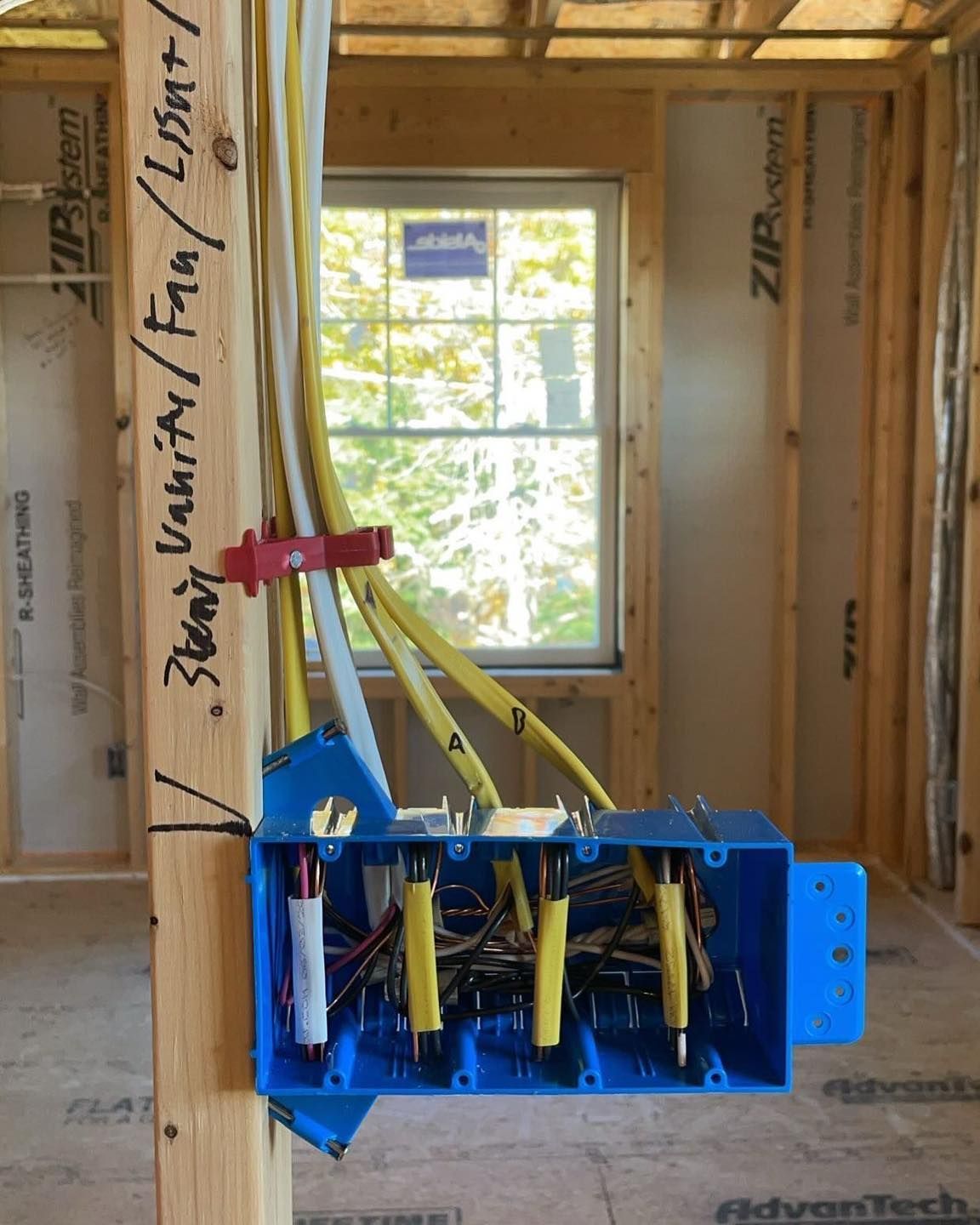 Blue electrical box in a framed room under construction, with yellow wires and a window in the background