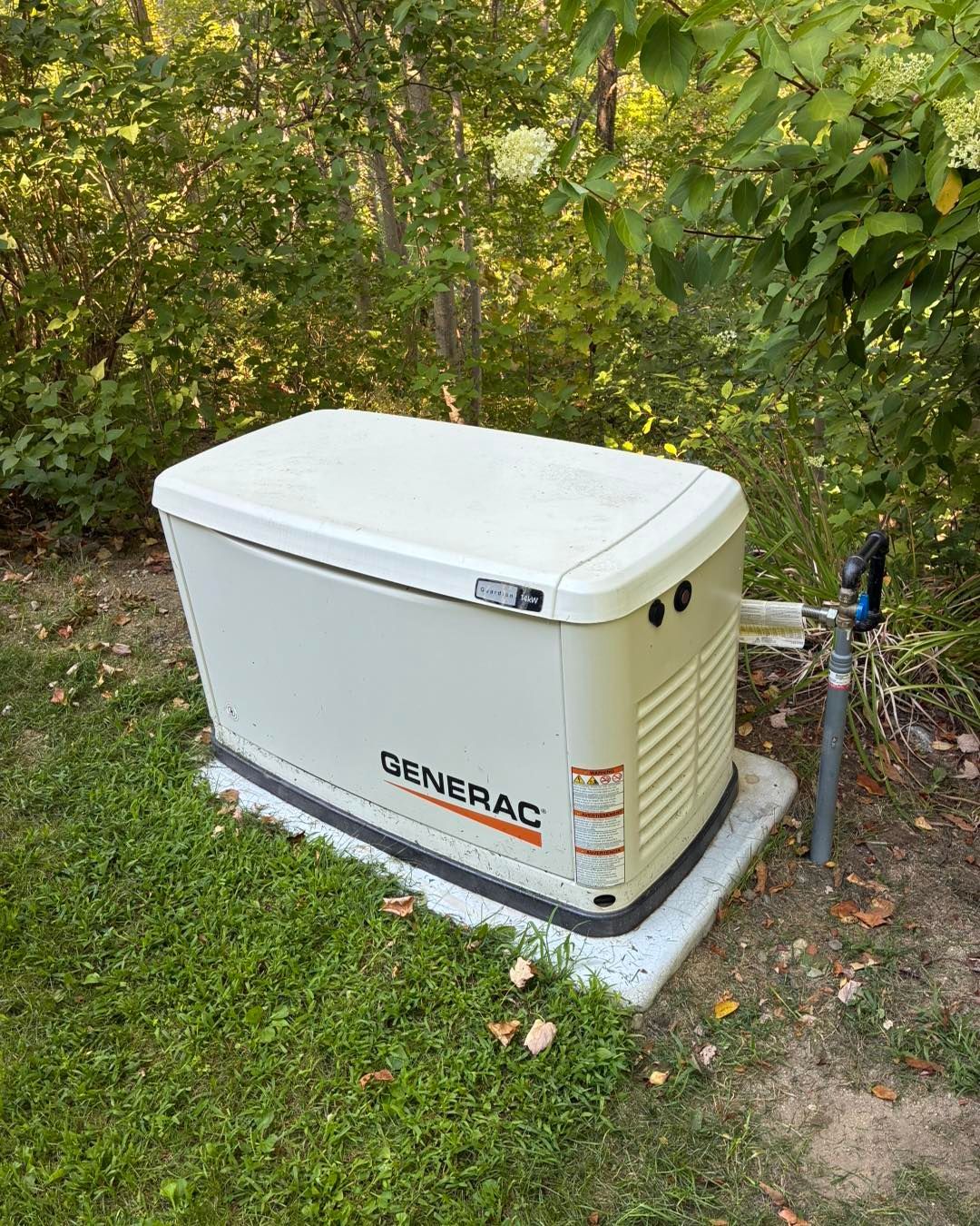 White Generac standby generator beside a house in a grassy yard, with trees in the background
