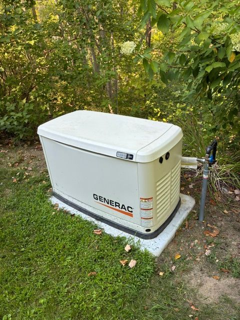 White GENERAC backup generator beside house, connected to utility lines, in a grassy yard.