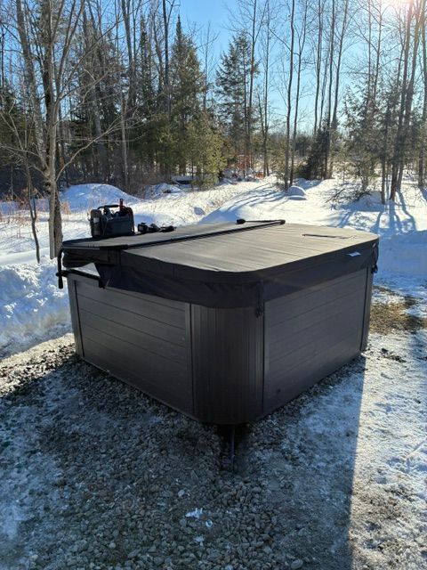 Covered outdoor hot tub in a snowy wooded yard with bare trees and sunlight