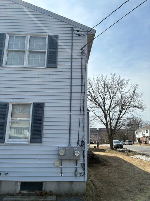 Side of a gray house with utility meters and a bare tree beside a street.