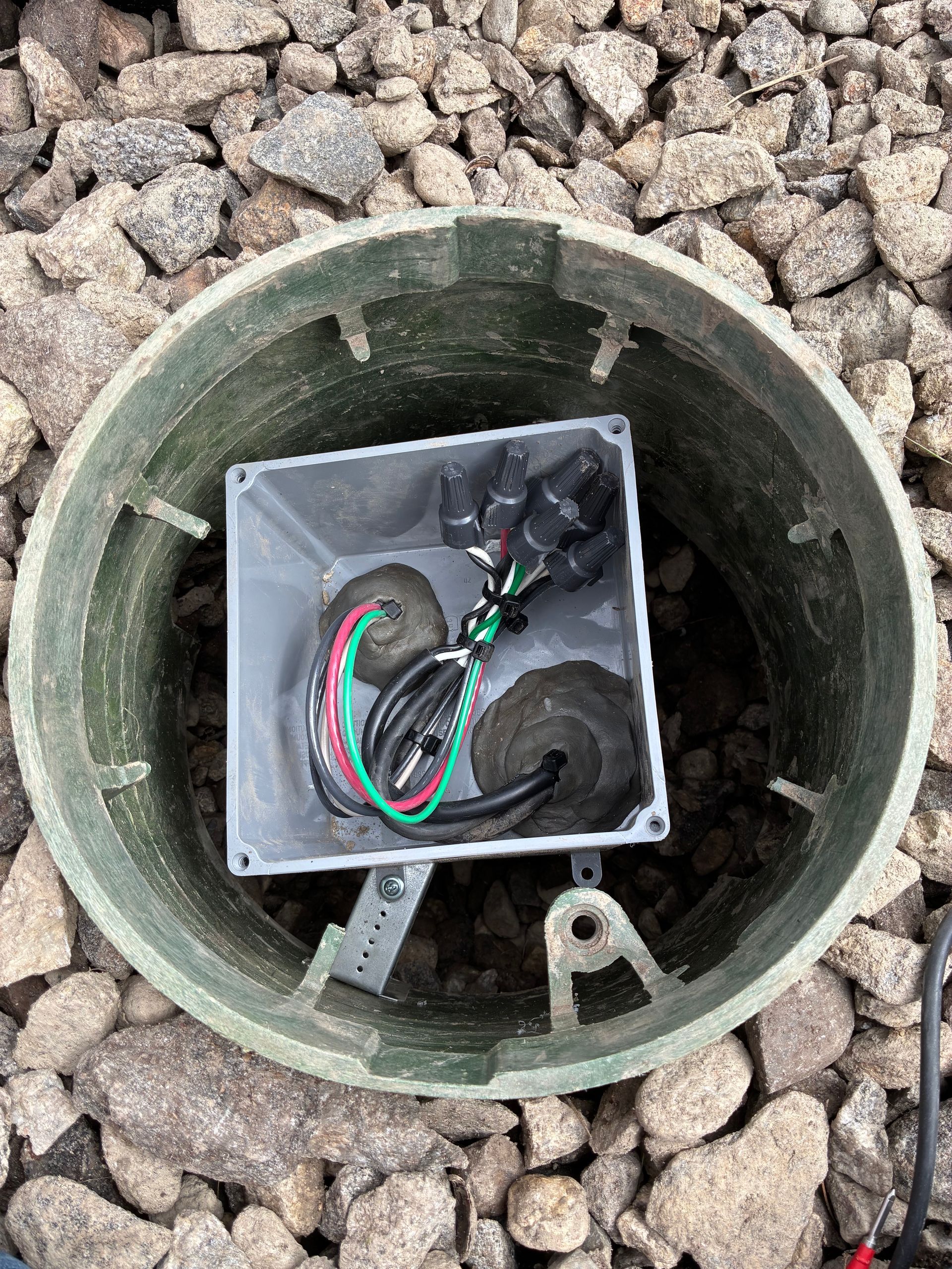 Open utility access hole with exposed electrical box and wires among gravel rocks