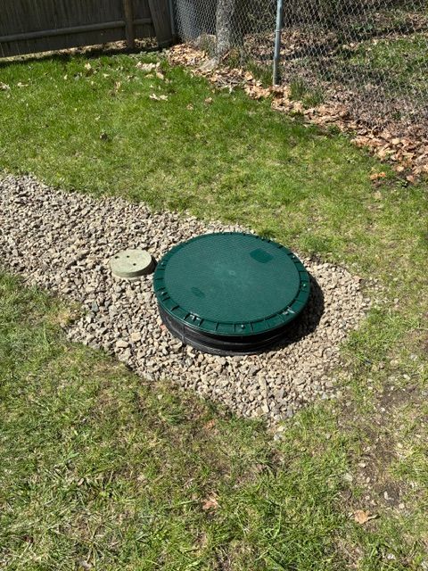 Green utility cover in a gravel patch beside a grassy yard and fence